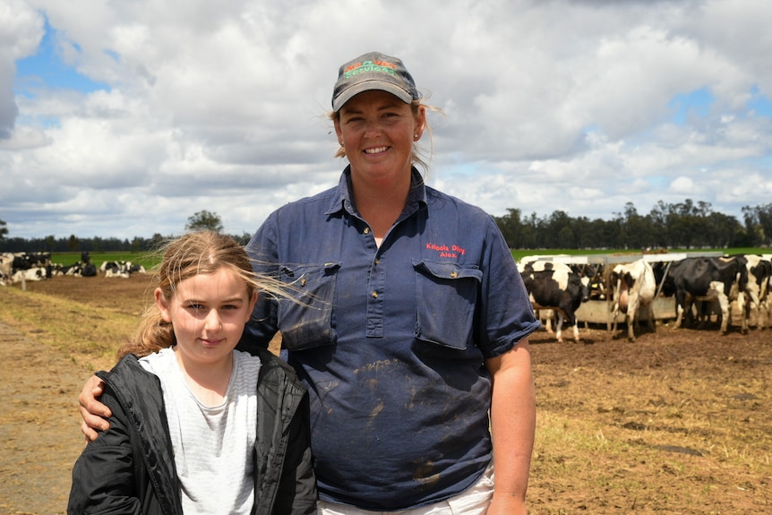 A woman in a blue shirt with a girl standing beside her. In the background are black and white cows.