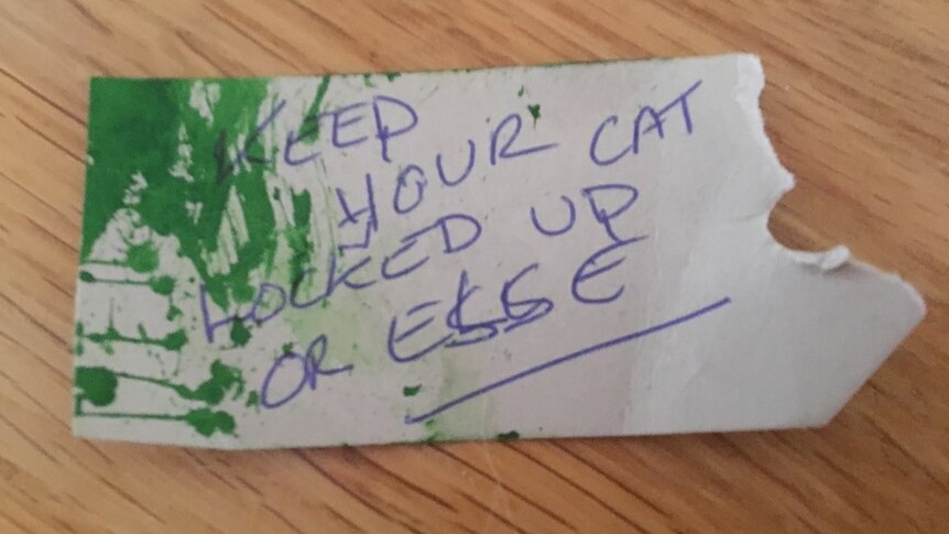 A torn piece of paper partially sprayed green has "keep your cat locked up or else" written in capital letters.