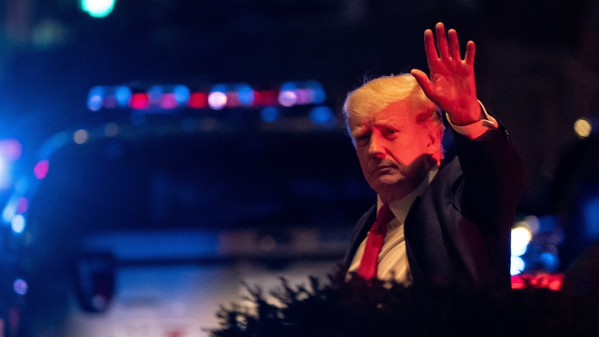 donald trump waves to camera lit up with red and blue emergency vehicle lights 