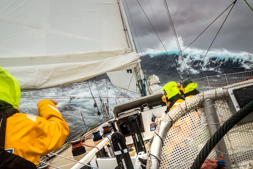 The front of a yacht heading into a big wave with three crew on board in yellow jackets. 