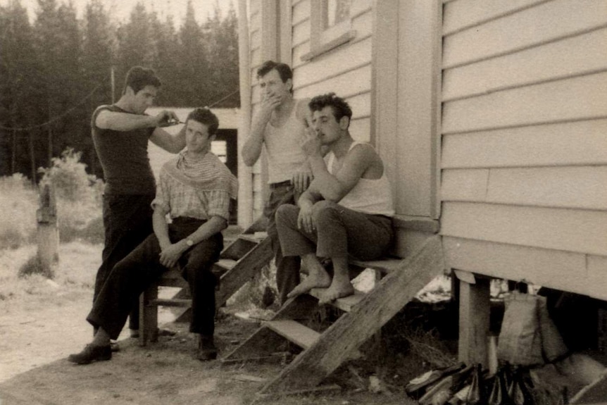 A black and white photograph of four young men gathering on the stairs of a wooden cabin, cutting each other's hair.