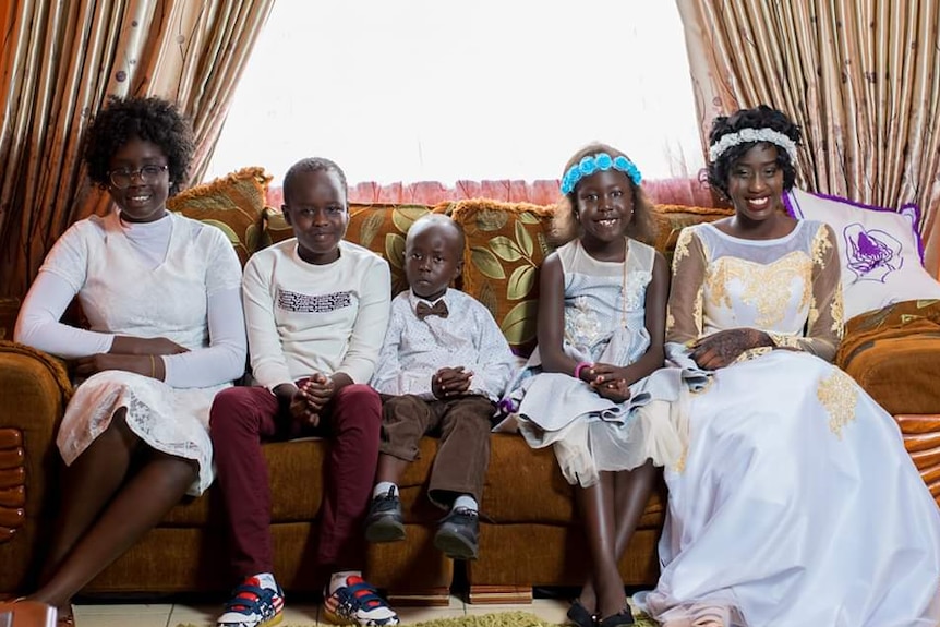 Five children sitting on an orange sofa in front of a window.