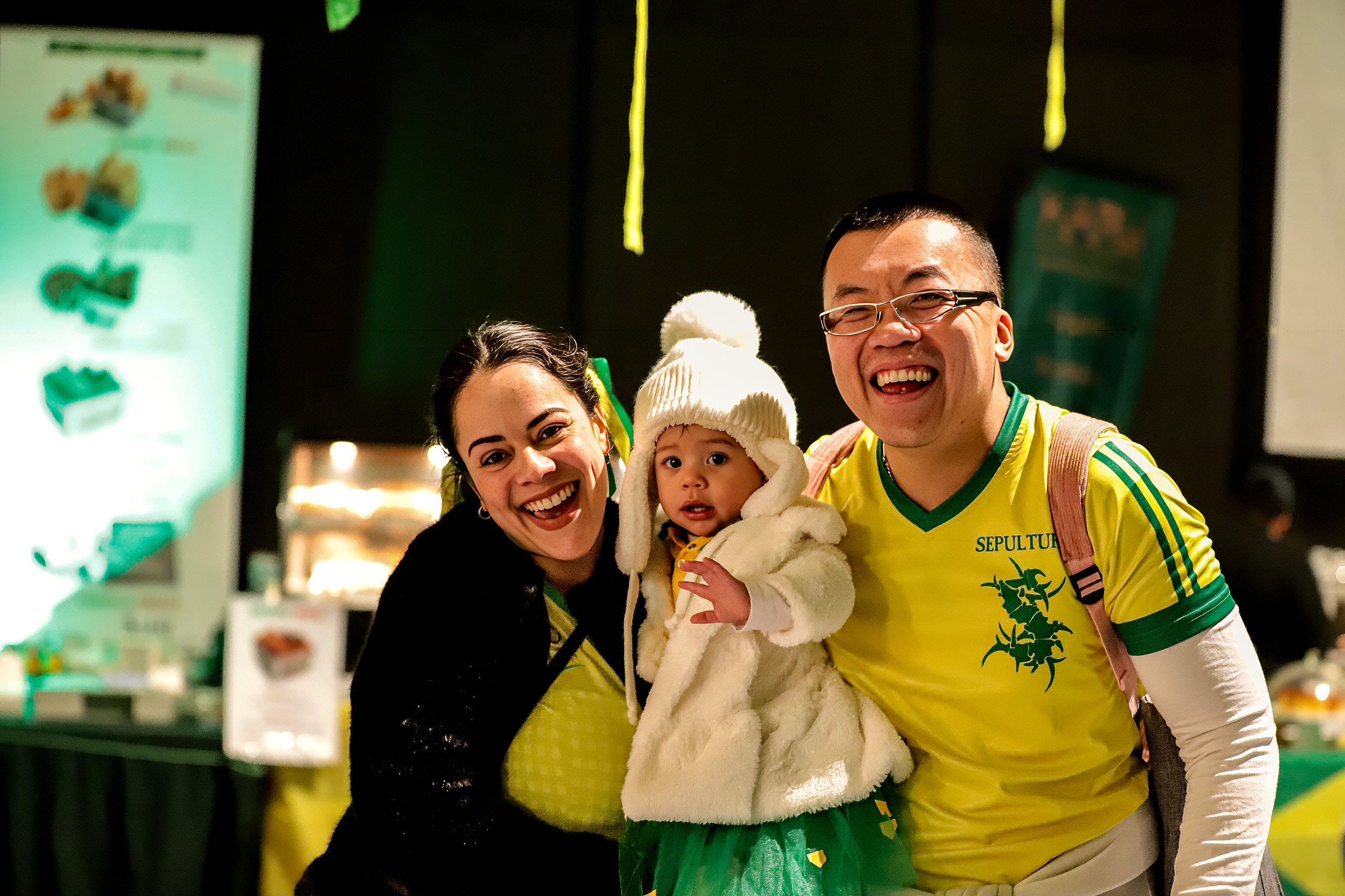 Man, woman and their young child, all wearing Brazillian yellow and green colours smile inside an event room