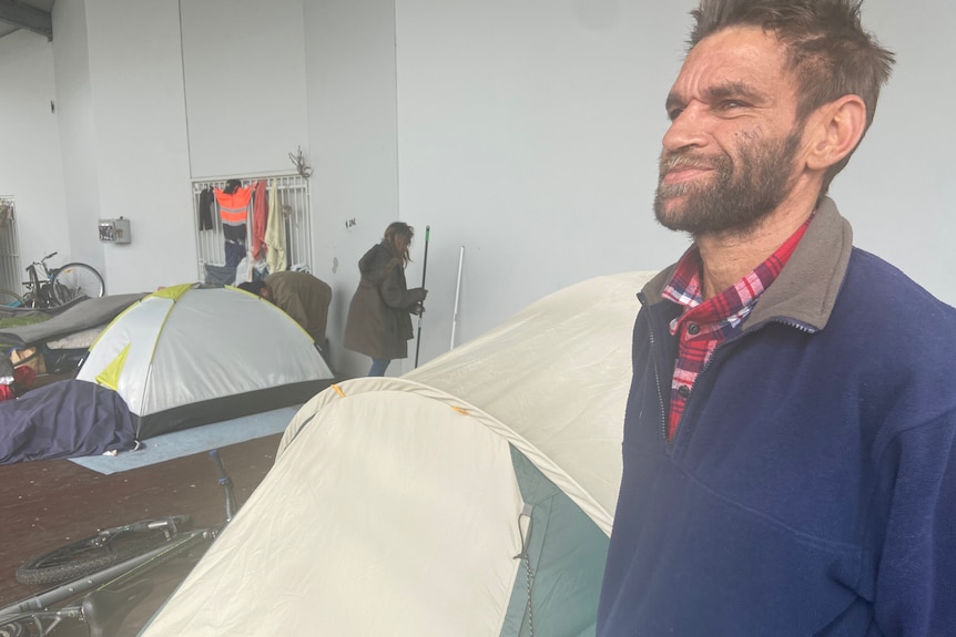 An Aboriginal man standing outside a tent in a sheltered area, in Bunbury., 