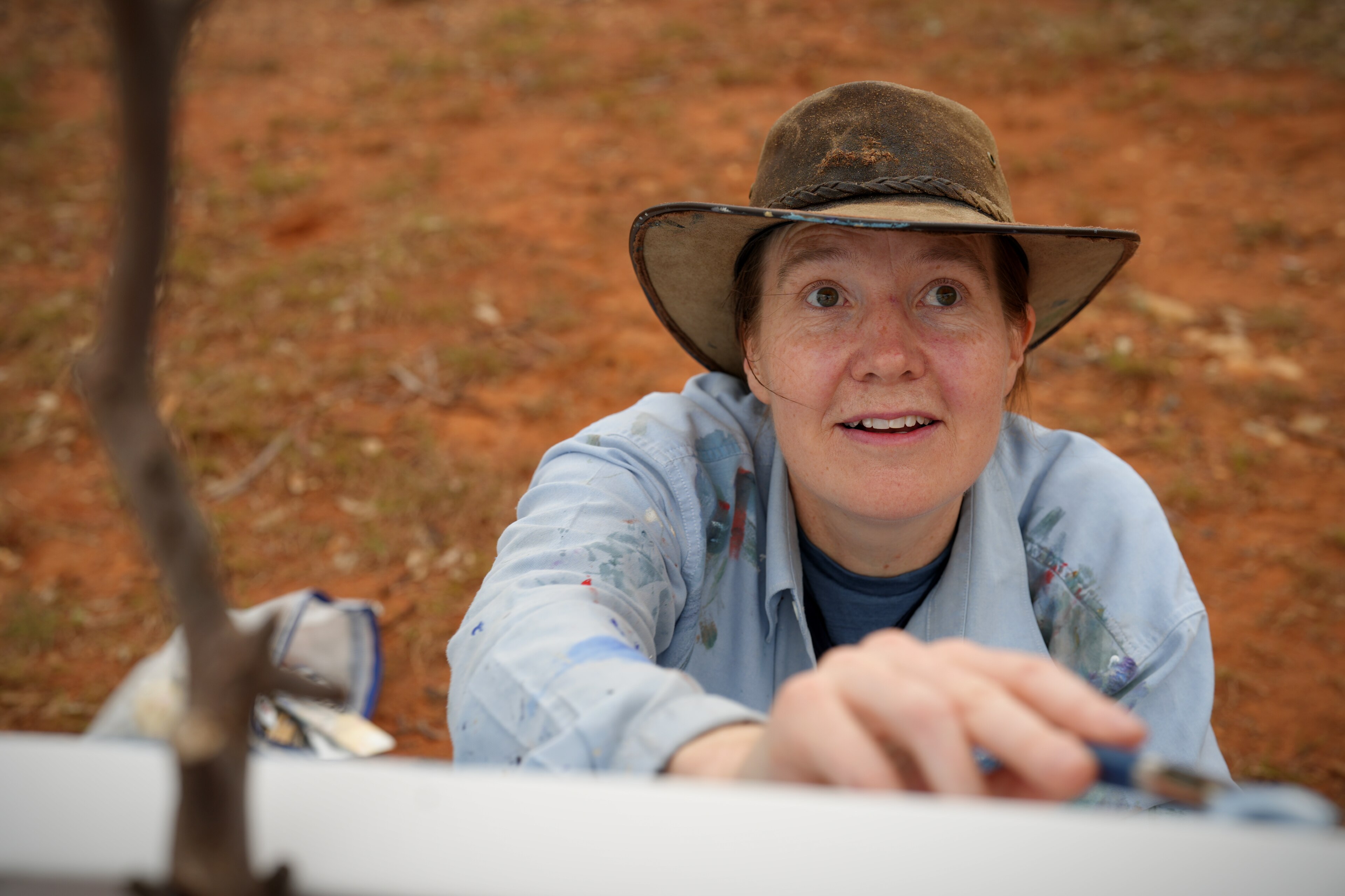 A woman in a paint-splattered button down shirt looks upward while painting on a canvas.