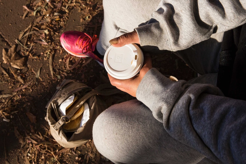 A homeless woman sits on a chair. On the ground her handbag is filled with pancakes to eat later that day.