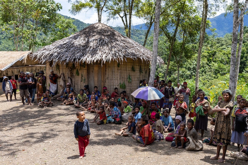 A line of women and children stand and sit in front of a hut with a straw roof, which is surrounded by trees.