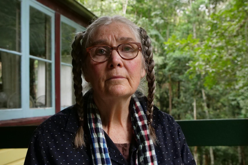 An older woman with glasses and two plats of brown and grey hair sits on her balcony surrounded by trees.