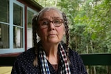 An older woman with glasses and two plats of brown and grey hair sits on her balcony surrounded by trees.