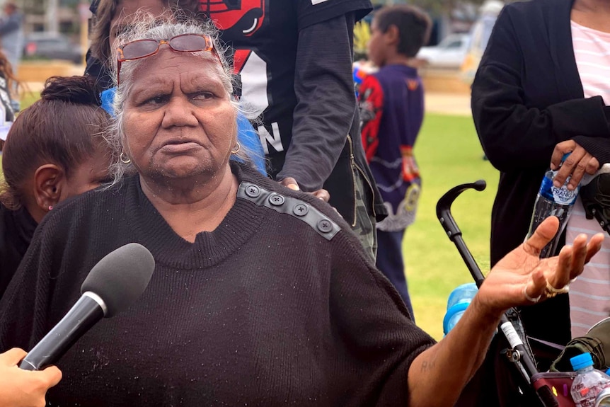 An older woman in a black jumper speaks into a microphone with a crowd of people behind her.