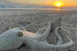 An octopus sculpture created from sand on a beach with a golden sunrise and blue sky in the background.