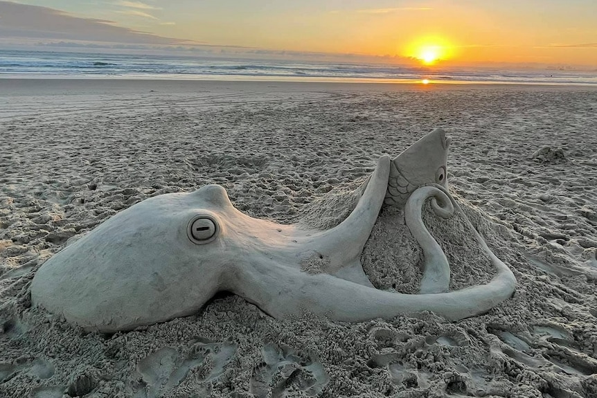 An octopus sculpture created from sand on a beach with a golden sunrise and blue sky in the background.