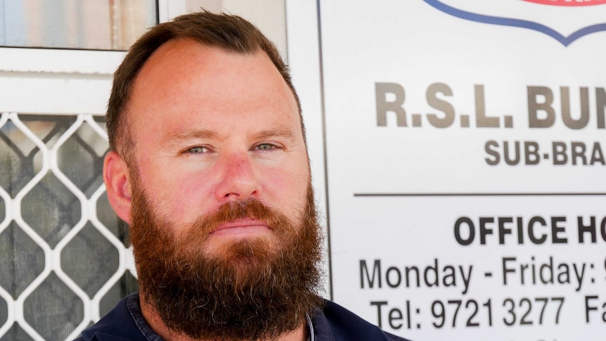 A man in his late 30s, with a beard, is standing in front of a white sign which says RSL Bunbury sub branch.