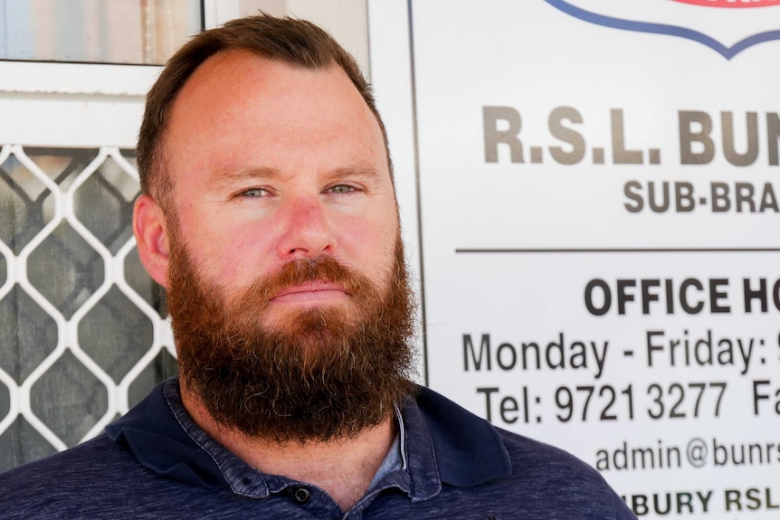 A man in his late 30s, with a beard, is standing in front of a white sign which says RSL Bunbury sub branch.