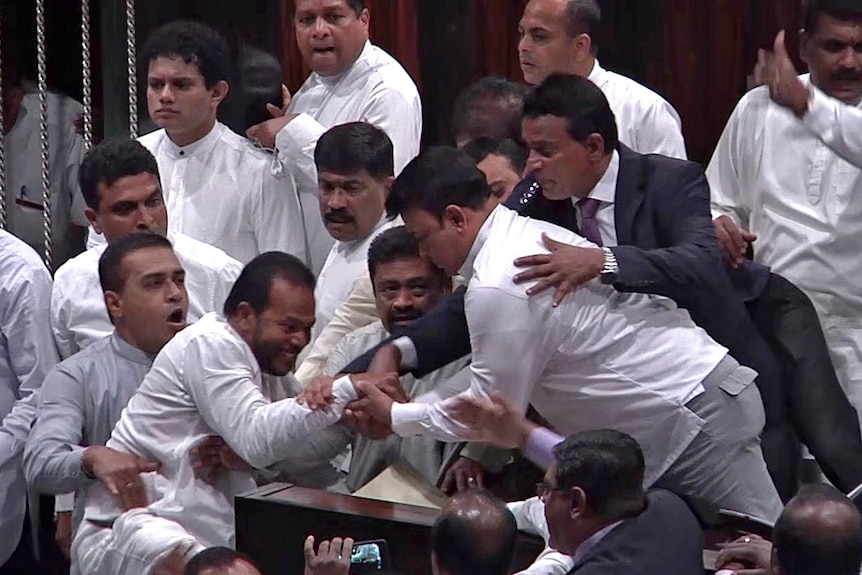 A group of men grasp each other's hands during a struggle watched by others.