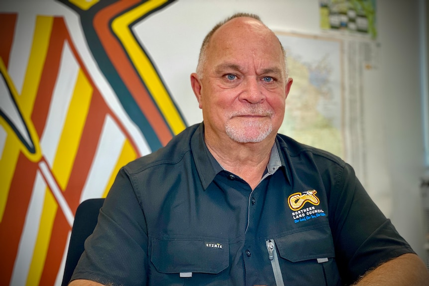 An older man with a neat white beard sits in an office with an Indigenous design on the wall.