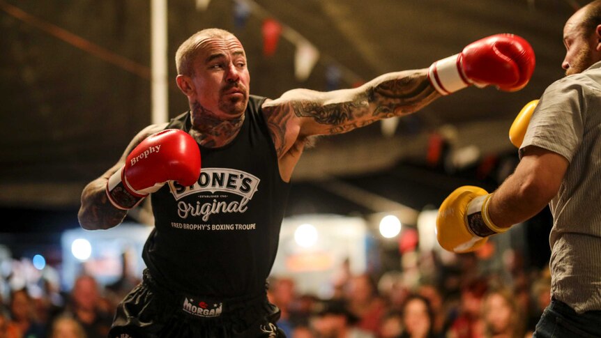 A man with tattooed arms and neck throws a punch at another man. He's wearing bright red boxing gloves with the word Brophy.