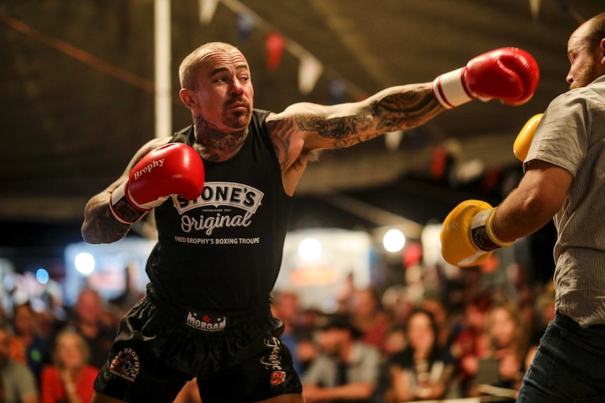 A man with tattooed arms and neck throws a punch at another man. He's wearing bright red boxing gloves with the word Brophy.