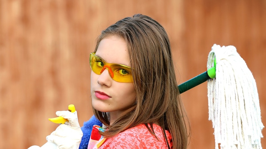 Woman wearing yellow protective glasses glares at camera, holding mop and cleaning spray