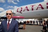 A balding man in a suit and tie stands outside next to a plane with Qatar Airways branding on it.