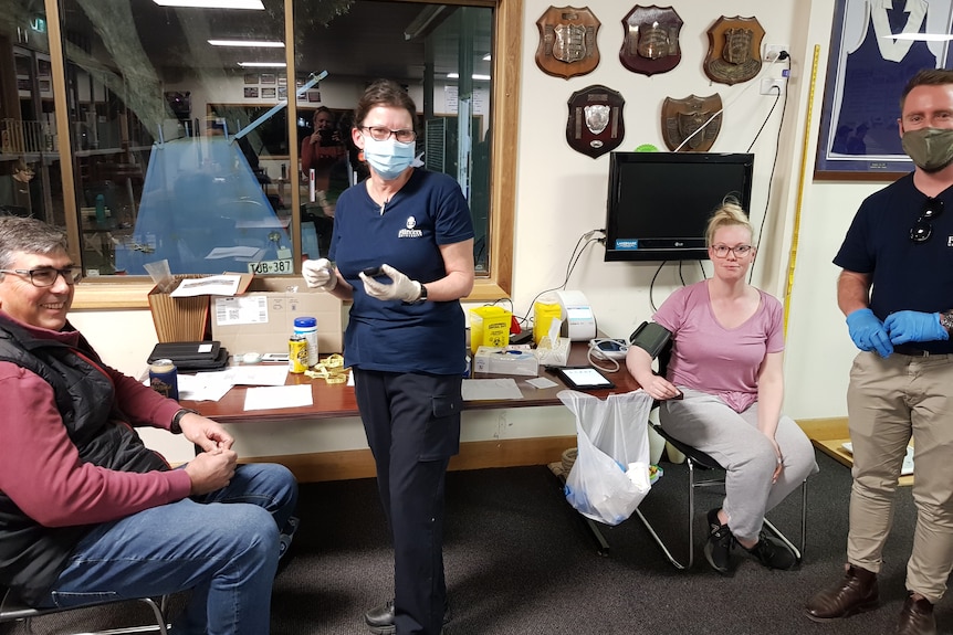 A man and woman in masks and gloves with two people sitting in chairs at a sports clubroom.