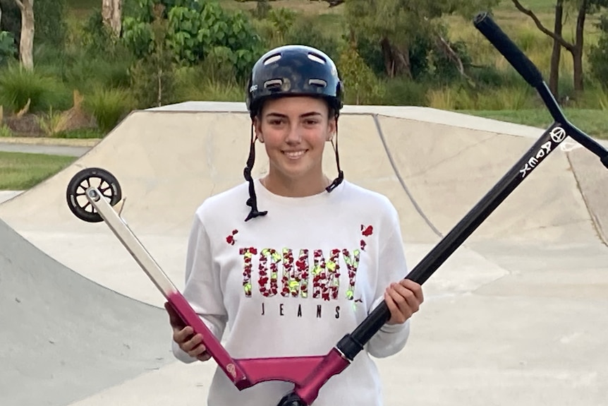 A smiling girl wearing a helmet and holding a pink and black scooter at a skate park