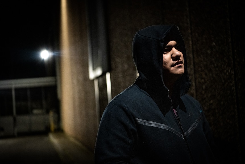 A man wearing a hoodie stands in a dark alley.