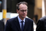 James Shipton sports slight smile as he walks along Sydney street wearing a dark suit, blue shirt and tie with black glasses.