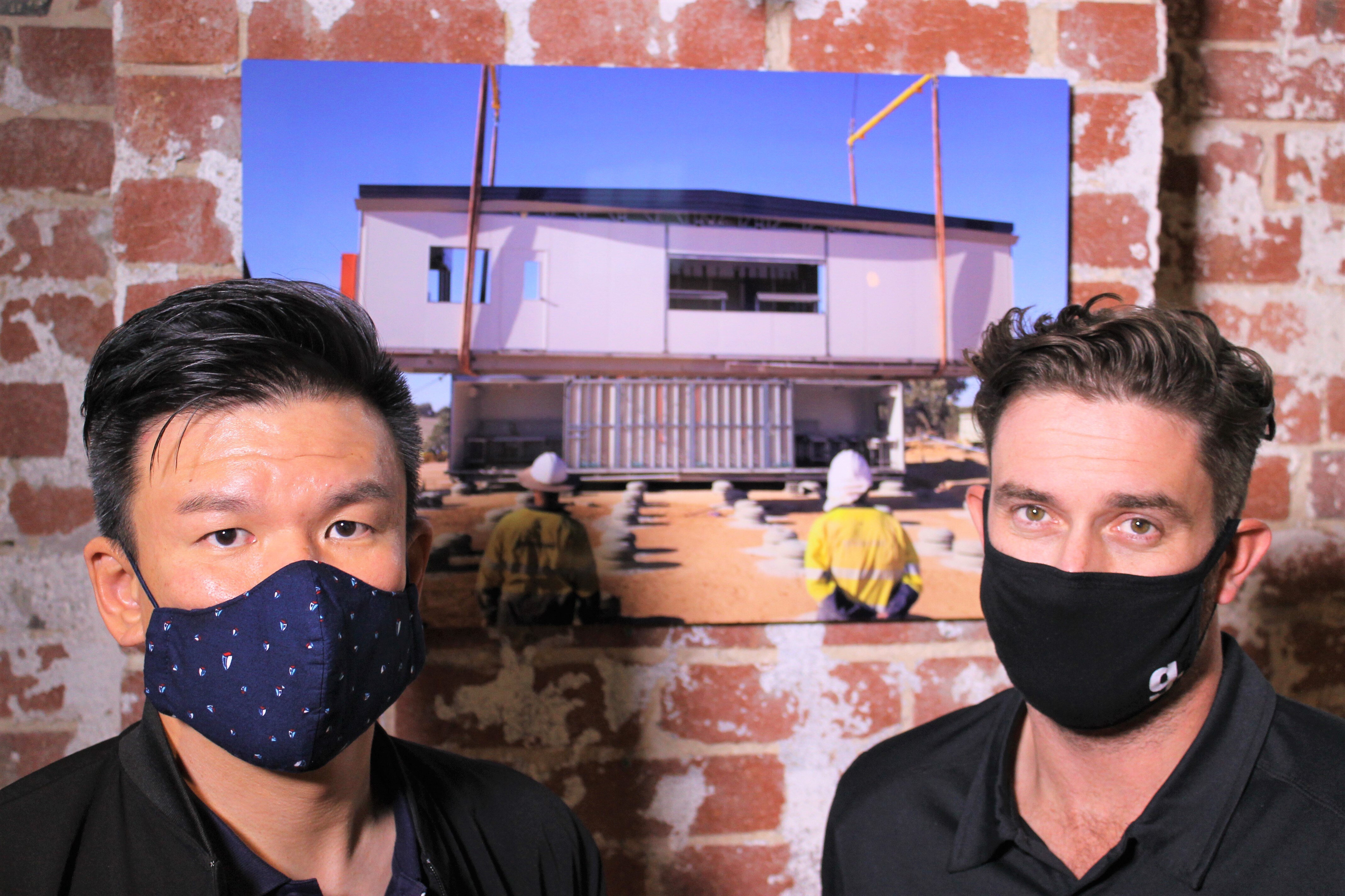 Two men wearing face masks stand in front of an exposed red brick wall with a photo frame in between them.