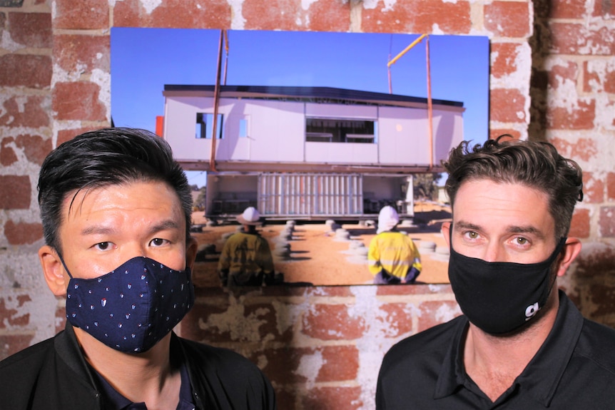 Dos hombres con máscaras faciales se paran frente a una pared de ladrillo rojo expuesta con un marco de fotos entre ellos.