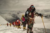 A line of mountaineers are seen in a single-file line down a white mountain peak.