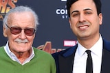 Stan Lee wearing a green jumper and his assistant Keya Morgan wearing a suit in front of an Avengers Infinity War backdrop