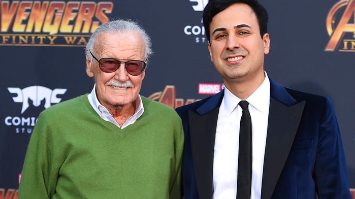 Stan Lee wearing a green jumper and his assistant Keya Morgan wearing a suit in front of an Avengers Infinity War backdrop.