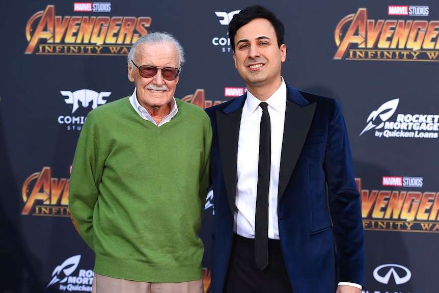 Stan Lee wearing a green jumper and his assistant Keya Morgan wearing a suit in front of an Avengers Infinity War backdrop.