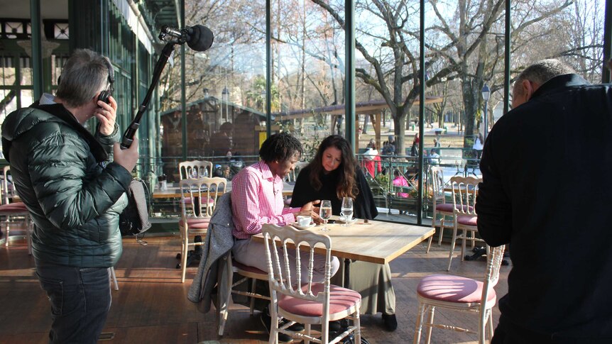 Man and woman sit at a cafe table talking as a television cameraman and soundman stand close by.
