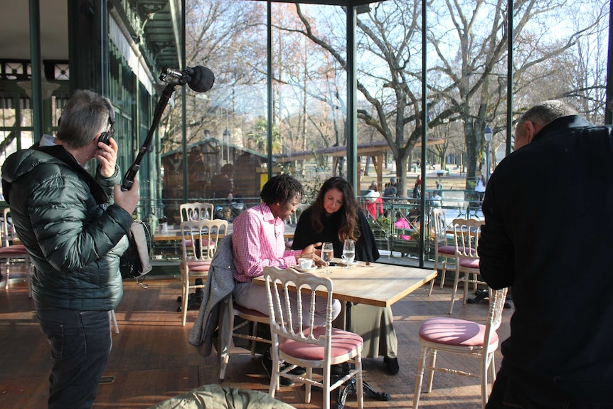 Man and woman sit at a cafe table talking as a television cameraman and soundman stand close by.