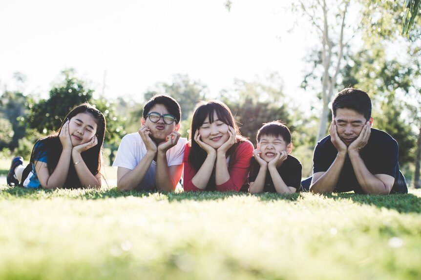 Parents and three kids lay in the grass. They have their heads in hands and are making fun silly faces.