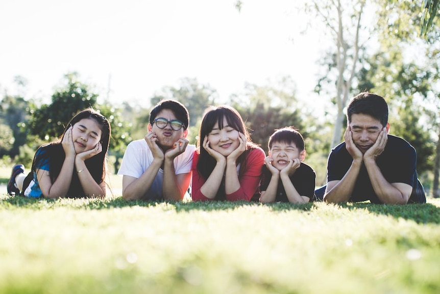 Parents and three kids lay in the grass. They have their heads in hands and are making fun silly faces.