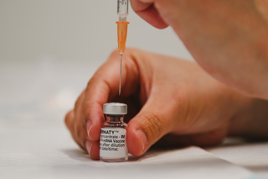 A close shot of a hand pressing a needle through the top of a vial of Pfizer vaccine.