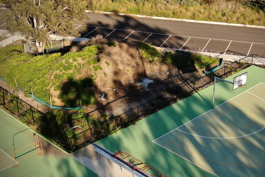 Netball court with dirt pile next to it, car park beyond, tree and grass