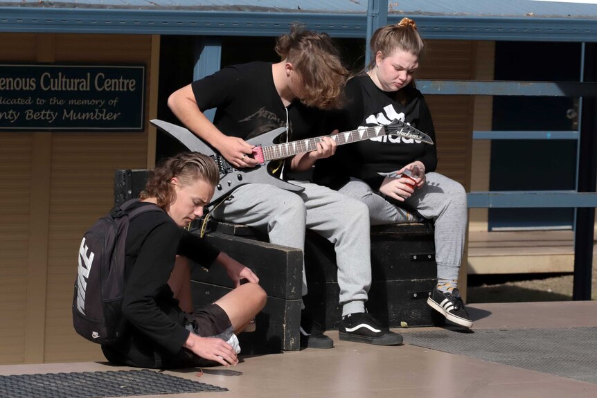 A group of three students sit in school yard, one plays a guitar