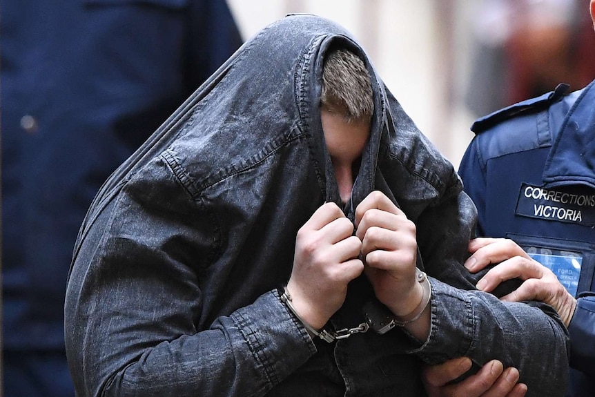 A man in handcuffs pulls a dark denim jacket over his head to cover his face. He is being led by police officers.