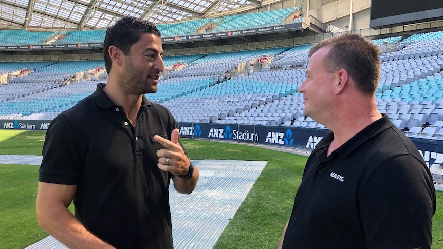 Anthony Minichiello stands talking with Dave Hendricks in front of rows of empty, blue stadium seats
