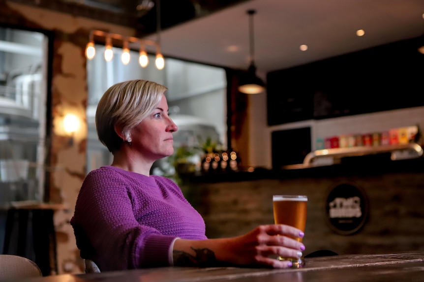 Woman with blonde hair and purple cardigan sits with beer in hand on wooden table inside brewery
