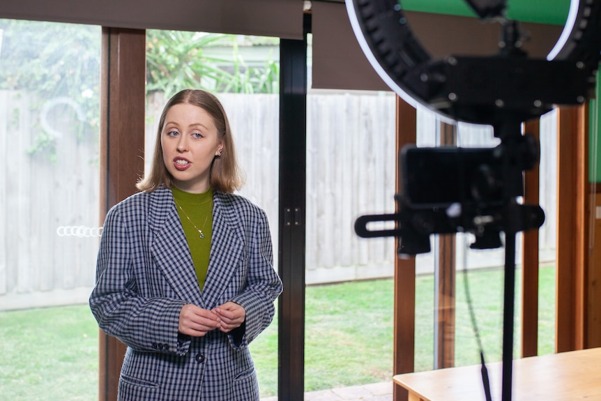 A young person stands and speaks in front of a ring light, which has a phone attached.