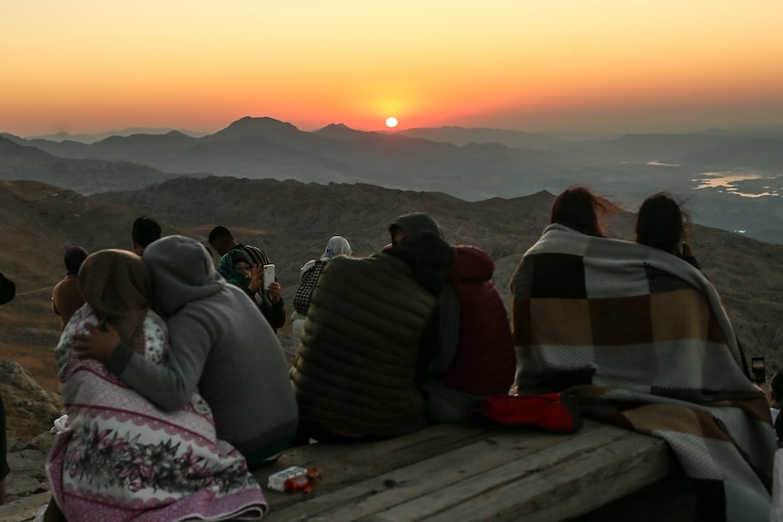 Couples huddle in warm clothing as they sit on a bench overlooking a spectacular view of a rocky landscape and orange sun.