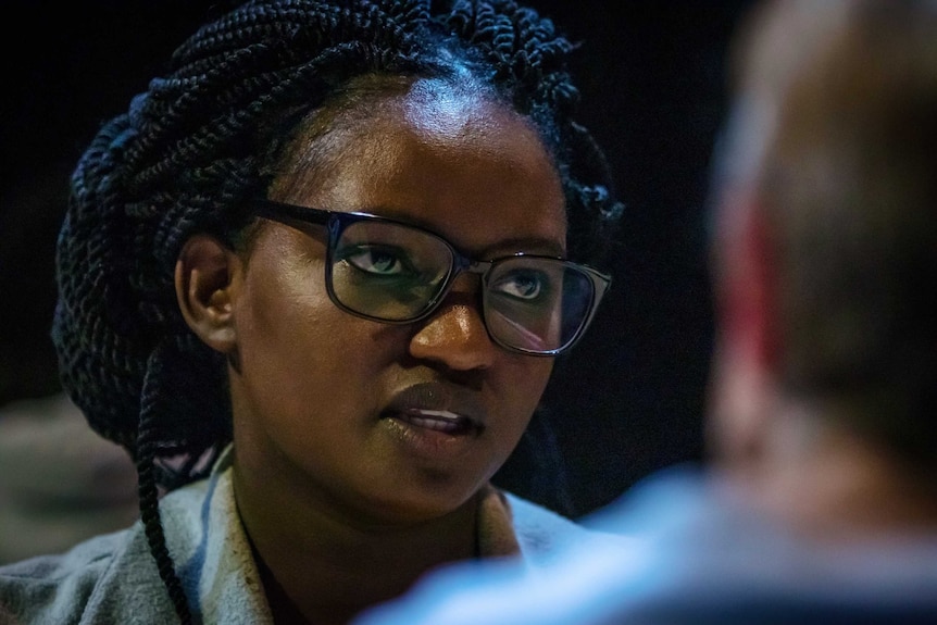 Chanceline Kakule looks to a person speaking who is seen from behind. Her braids are tied back and she wears black glasses.