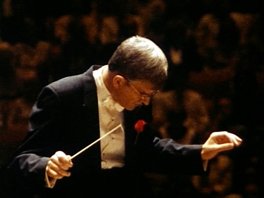 An orchestra conductor wearing a suit and white bow tie looks down. One hand holds a baton, the other is indicating the tempo.