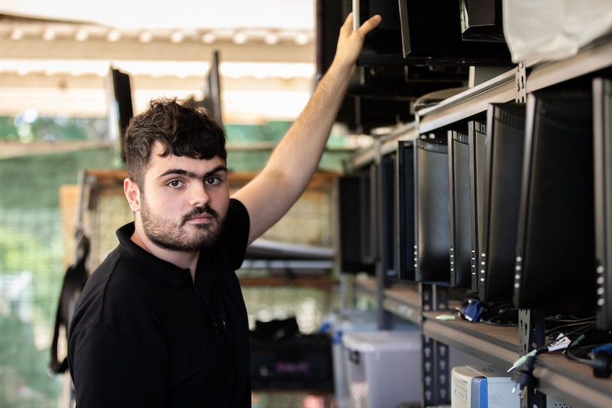 A man in a black shirt holding a computer monitor next to a rack full of monitors.