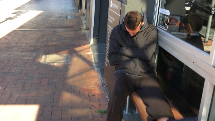 A man in a worn coat sits huddled against a building with his arms tightly around himself 